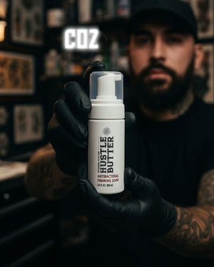 Hustle Butter Antibacterial Foaming Soap 7oz 207ml bottle held by tattoo artist wearing gloves in studio setting, professional tattoo cleansing soap used during tattoo sessions, available at COZ Tattoo Supplies