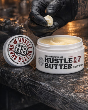 Open jar of Hustle Butter Tattoo Balm Deluxe 5 oz on tattoo workstation, artist wearing black gloves holding balm, available at COZ Tattoo Supplies
