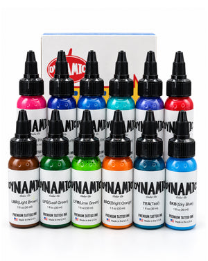 Twelve bottles of Dynamite Premium Tattoo Ink arranged in two rows in front of a branded box, showing a full range of colors including pink, blue, red, green, orange, and brown on a white background.