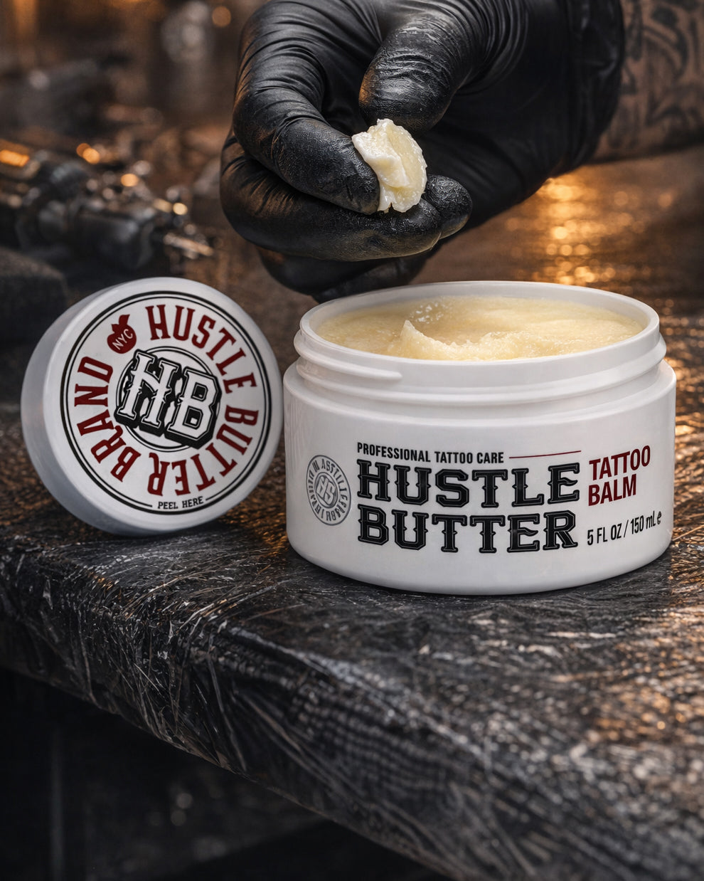 Open jar of Hustle Butter Tattoo Balm Deluxe 5 oz on tattoo workstation, artist wearing black gloves holding balm, available at COZ Tattoo Supplies