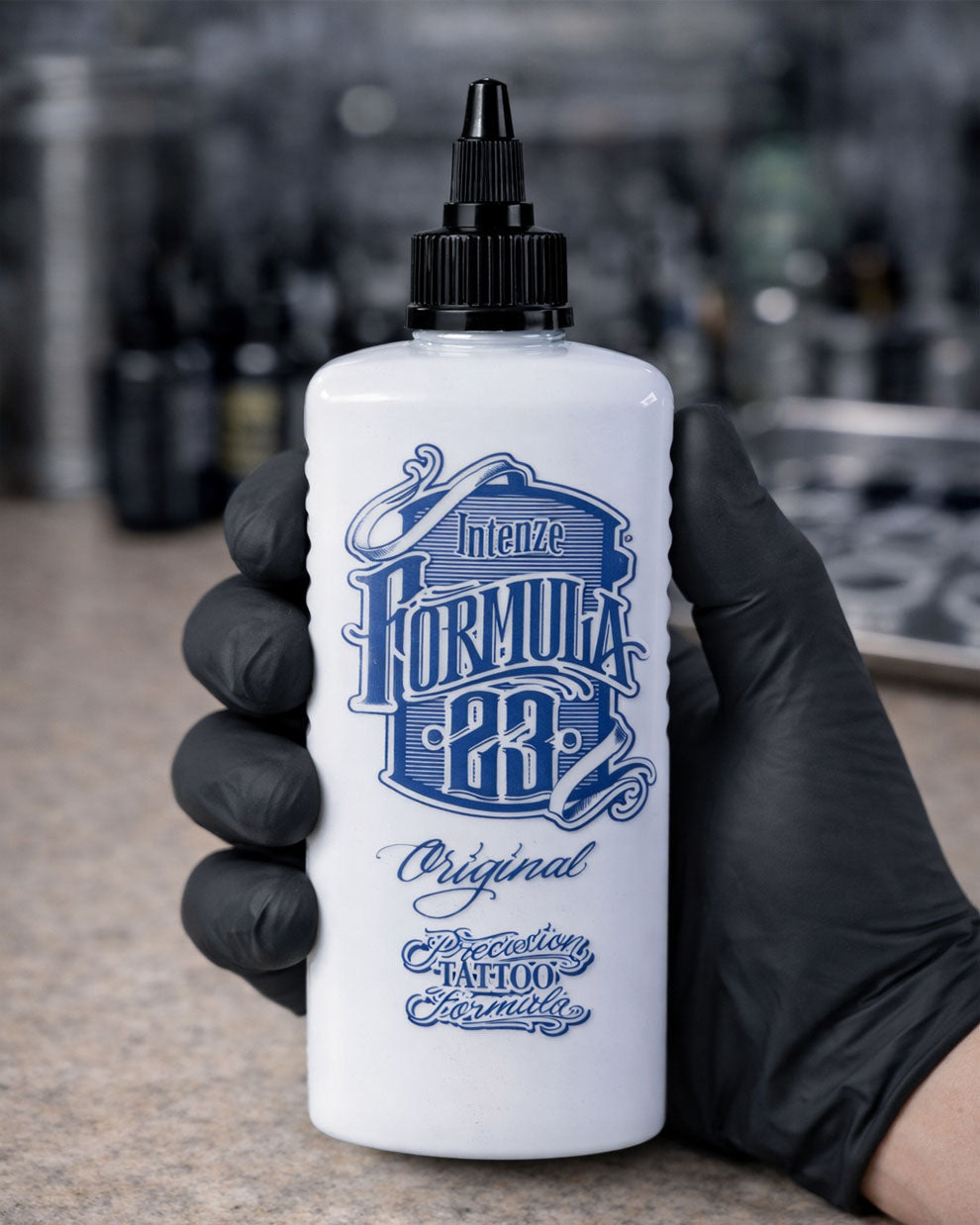 White bottle of Intenze Formula 23 tattoo ink held by a gloved hand with a blurred background.