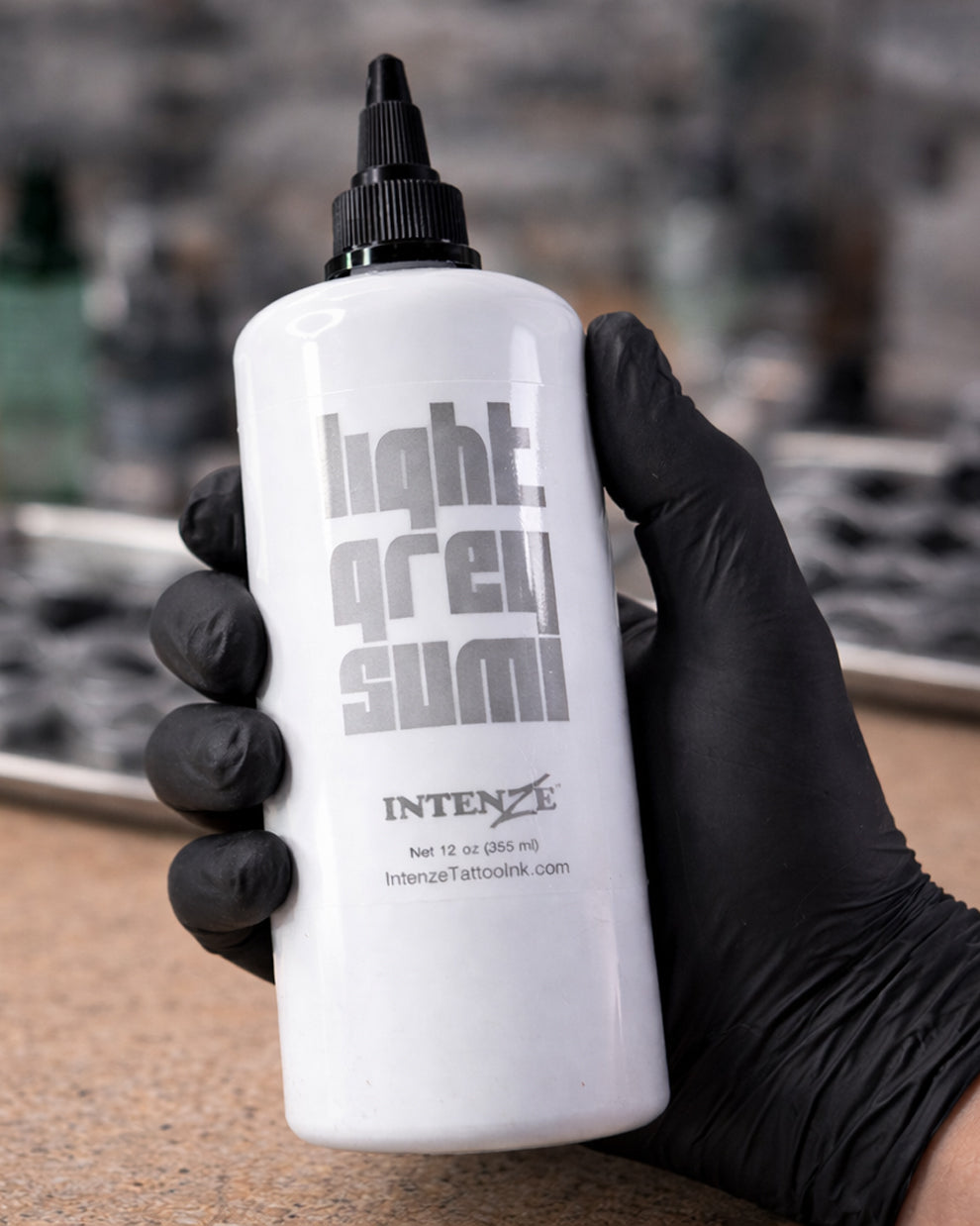 Intenze Light Grey Sumi tattoo ink bottle held by a tattoo artist wearing black gloves, professional studio photo on clean background, Coz Tattoo Supplies